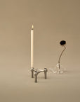 A minimalist silver metal candle holder with a single lit white taper candle, styled next to a clear glass vase holding a dried black flower, set against a warm beige background.
