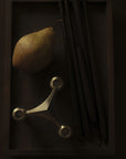 A dark wooden tray holds a brown pear, a metallic three-spoke candle stamd, and several thin black rods against a very dark background.