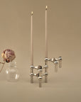 A polished silver geometric candle holder with two tall lit taper candles, styled next to a small clear glass vase holding a dried pink rose against a warm beige background.