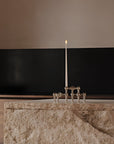 A silver-toned chrome metal candle holder with a single lit white candle, set on a textured stone countertop against a dark wall and neutral upper background.