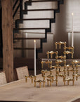 A gold-toned geometric candle holder sculpture arranged on a wooden dining table, with several white taper candles placed around it in a softly lit interior setting with a staircase in the background.