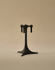 A matte black metal jewelry display stand with a three-prong top and a wide, stable base, set against a solid warm beige background.