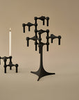 A black sculptural multi-arm candelabra and matching candlestick holder with a white candle, photographed on a warm beige studio background.