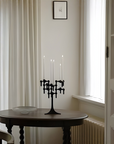 A black metal candelabra with four candle holders stands on a round dark wood table in a softly lit room with cream curtains, a window, and a small framed picture on the wall.