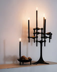A black metal multi-arm candle holder with four tall candle sleeves, styled on a shelf next to a matching black candlestick and a small bowl of stones against a warm off-white wall.