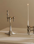A polished silver-toned metal candlestick holder set on a draped cream tablecloth against a warm beige background. One holder is empty, and the other stands with a single lit white taper candle.