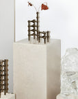 A set of small bronze-finished metal garden stakes or trellis pieces displayed on a white stone pedestal, with a clear textured glass vase in the foreground and a white wall behind.