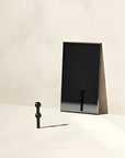 A black metal wall-mounted triangular shelf or hook stand with a small matching black hanging peg, photographed against a clean off-white wall and surface.