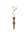A decorative brass-toned metal vase with a vertical cylindrical body, two stacked round bead-like spheres, and three dried fluffy stems inserted at the top, shown on a plain white background.