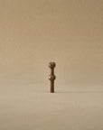A small bronze metal hardware piece with a vertical shaft and two rounded decorative ends, centered against a plain warm beige background.