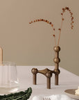 A bronze-finished sculptural vase cum candle holder with a geometric, tiered design holding dried brown stems, styled on a white table next to a glass and a plate of food.