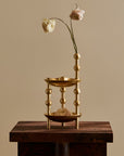 A gold-toned two-tier decorative stand with bead-like vertical accents, displayed on a dark wooden pedestal against a warm beige background. The top tier holds a dried white flower, and the bottom tier holds a dried beige flower.