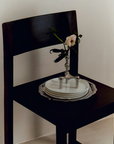 A dark wooden chair in a minimalist room setting, styled with a stack of white plates on a silver pedestal stand holding a single pale pink flower.