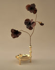 A decorative brass vase and candle holder with three dark brown flower-like petals on curved stems, set against a plain beige background.