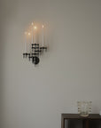 A black metal multi-arm wall sconce mounted on a light gray wall, styled above a dark wooden console table with a clear glass vase and a book.