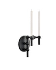 A modern wall-mounted candle sconce in matte black, featuring a round backplate and two slim white candle-style arms, shown against a plain white background.
