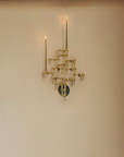 A modern brass wall-mounted candelabra with two tall candle-style arms and multiple tiered branches, shown lit against a warm beige wall.