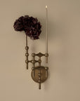 An ornate wall-mounted candle holder with a bronze-finished metal frame, featuring one arm holding a tall white taper candle and the other holding a dark burgundy dried flower, set against a plain beige wall.