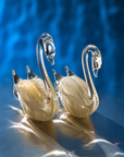 Two decorative crystal swan figurines with cream-colored feathers and gold accents, set against a shimmering blue background.