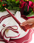 A hand holds a dark red led taper candle in front of a matching dark red and white place setting, with a folded white napkin, scalloped placemat, and festive green garland in the background.