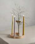 A minimalist four-arm candle holder made of light natural wood, holding four slim led taper candles in pastel yellow, lavender, and mustard tones, with dried grasses in a small pot, set on a white tablecloth.