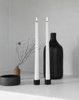 Two white led taper candles in black holders on a white shelf, styled with a matte black vase and a small dried branch vase against a light gray wall.