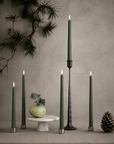 Five dark green led slim taper candles with silver and black holders are arranged on a neutral beige surface against a textured beige wall, styled with a pine branch, a small white pedestal, a green bulb, and a pinecone.