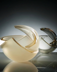 Two sculptural glass vases with abstract crescent-shaped forms, one in glossy cream and the other in smoky translucent gray, displayed on a reflective surface against a dark gradient background.
