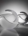 Two abstract sculptural glass art objects with curved, broken-edge bands, one frosted white and one transparent smoky gray, displayed on a reflective surface against a dark gray gradient background.