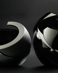 Two abstract sculptural black glass objects with glossy reflective silver curved bands, photographed against a dark charcoal studio background.