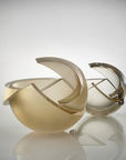 Two abstract sculptural glass art objects with curved, broken-edge forms, one in brushed gold and one in smoky translucent gray, displayed on a reflective surface against a light gray studio background.