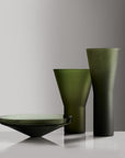 Three translucent dark green glass vessels on a reflective white surface against a gray gradient background: a shallow wide bowl, a medium conical vase, and a tall flared vase.