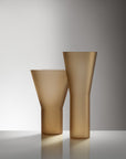 Two frosted glass vases in a warm champagne-gold tone, one with a flared top and one with a tapered cylindrical shape, standing on a reflective surface against a gray gradient background.