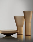 Three matte glass tableware pieces, including a shallow bowl and two tapered vases, arranged on a reflective surface against a neutral gray background.
