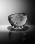 A glass art object with a textured, bubbled interior, shown on a reflective dark surface against a gray gradient background.