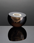 A round glass art object with a glossy dark amber-brown body, a clear top rim, and a textured metallic gold interior, shown on a reflective gray surface against a dark gray background.