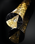 A close-up of a glossy cylindrical sculptural vase and dense metallic gold glitter or foil pattern, photographed on a dark reflective surface.
