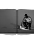Assouline Louis Vuitton: Virgil Abloh Classic Cartoon Cover coffee table book displaying Grey Image of Virgil Abloh using Apple Mac Laptop on a white back ground available at Spacio India for luxury home decor accessories collection of Fashion Coffee Table Books.