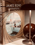 Assouline James Bond: Destinations coffee table book beside table lamp on console available at Spacio India for luxury home decor accessories collection of Travel Coffee Table Books.