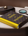 A Louis Vuitton book set displayed on a light stone table, featuring a dark brown checkered hardcover edition with gold lettering and a bright yellow companion book. A dark brown oval box with a gold lid and a magnifying glass are arranged nearby.