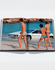 An open hardcover book laid flat on a light gray background, showing a printed beach scene with three people in swimwear walking beside a white car on the sand, with blue water and rocky shoreline across the spread.
