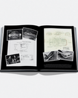 An open hardcover book laid flat on a light gray background, showing two printed pages with black-and-white photographs, handwritten notes, and technical drawings on a black page background.