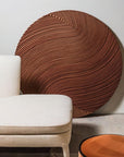 Round brown sculptural wall art on a light gray wall next to a white chair a round wooden wall art piece