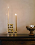 A brass candelabra with four lit taper candles displayed on a dark wooden console table against a warm beige plaster wall, with a decorative woven bowl to the right.