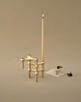 A gold-toned geometric candle holder with a tall unlit taper candle, photographed on a warm beige studio background. A small white dish with dark contents and a small dark object sit to the right on a folded white paper.