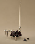 A chrome finish candle holder with a tall white taper candle and a bowl of dark burgundy rose petals, set against a warm beige background.