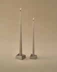 Two tall taper candles in square metal holders, lit and standing against a warm beige studio background.