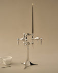 A polished metal candelabra with a tall central candle holder and multiple smaller candle arms, shown on a warm beige studio background. A small white cup and spoon sit to the lower left as styling props.