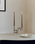 A polished silver metal candelabra with two tall black taper candles lit, displayed on a white table in a minimalist interior setting.