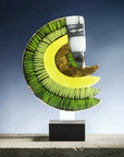 Abstract crescent moon sculpture with green and yellow segments, a white and gray face motif, and gold leaf accents, displayed on a black rectangular base against a blue gradient background.